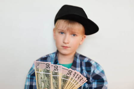 A photo of handsome stylish liitle rich child dressed in black cap and modern shirt holding dollars in his hands. Young businessman showing money isolated on white background. Selective focus.の写真素材
