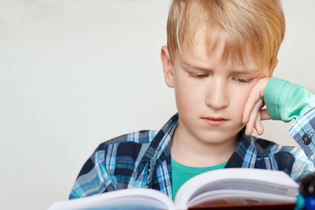 Book, school, kid. Little schoolboy with blond hair dressed in checked shirt siting and looking into the book having tired expression. A little boy preparing for the lessons reading a bookの写真素材