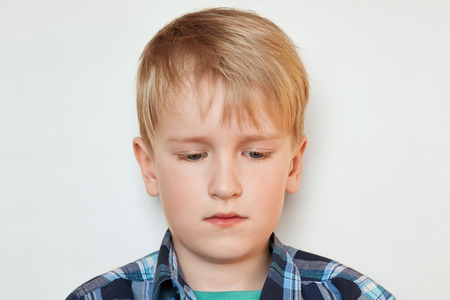 A close-up portrait of handsome little boy with fair hair and blue eyes dressed in checked shirt having sad expression looking down isolated over white background. People and emotions concept.の写真素材
