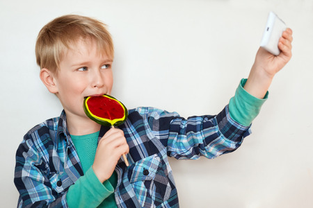 Adorable little boy licking big colorful lollipop and making selfie by his smart phone isolated on white background. Fashionable kid having fun tasting sweet candy and making photosの写真素材