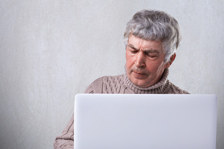 A mature man with gray hair siting in front of his laptop having dissatisfied expression and wrinkles on his face isolated over white background. People, emotions, technology and lifestyle concept.の写真素材