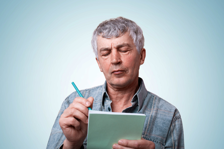 A portrait of mature man with gray hair wearing jean stylish shirt holding notebook and pen in his hands making some notes having serious expression while standing over blue background.の写真素材