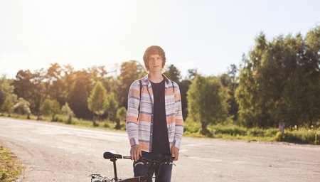 Active summer holidays. Portrait of young biker dessed in casual shirt looking seriously into camera enjoying ride outdoors by his bicycle. Teenager having rest during his morning cycle rideの写真素材