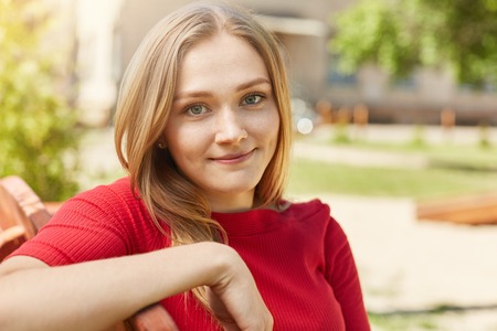 Pretty blue-eyed woman with fair hair relaxing outdoors while sitting at bench looking directly into camera rejoicing having her summer holidays. Cute female resting in open air admiring natureの写真素材