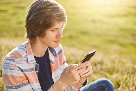 Stylish teenager sitting at green garss outdoors holding smart phone in his hands typing messages or reading newsfeed. Seriois boy using modern gadget for entertainment playing different games onlineの写真素材