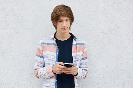 Young hipster guy with trendy haircut having concentrated look in his smart phone downloading pictures online using free internet conncetion. Schoolboy using cell phone surfing social networksの写真素材