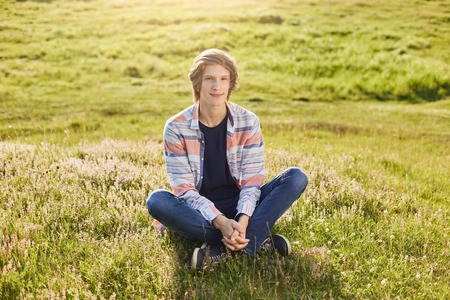 Candid shot of handsome teenager boy sitting crossed legs at green grass of field looking directly into camera with pleased expression keeping his hands together while having good rest outdoorsの写真素材