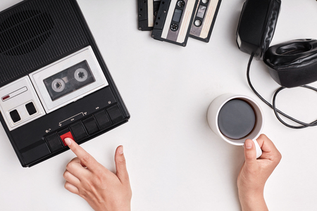 Top view of retro tape recorder, cassettes and headpnones lying on white surface. Woman`s hands switcing on old tape recorder listening to music holding cup of coffee in hand remembering somethingの写真素材