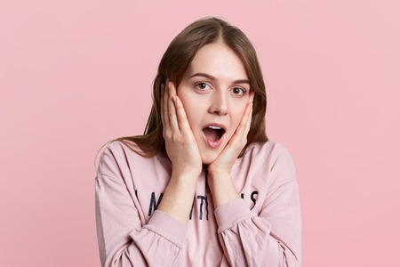 Headshot of shocked beautiful woman keeps hands on cheeks, looks in terror at camera, wears casual...