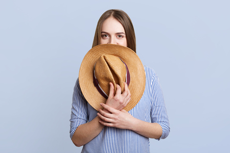 Pretty mysterious woman in blue clothes hides under straw hat, poses against blue studio background. Fashionable beautiful young cute female demonstrates her new purchases after going shoppingの写真素材