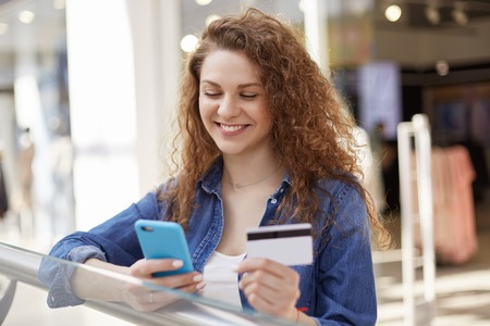 Positive female model dials number of credit card on smart phone, checks her bank account, makes...
