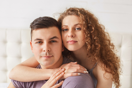 Shot of pleasant looking young female hugs her handsome boyfriend, pose together against domestic interior, spend day off at home, dressed casually. People, family, lifestyle, mutual love conceptの写真素材