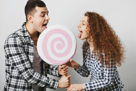 Two female and male friends lick big lollipop together, like sweets and candies, being sweety tooth, isolated over white studio background. Pretty curly woman and her boyfriend with sweet candyの写真素材