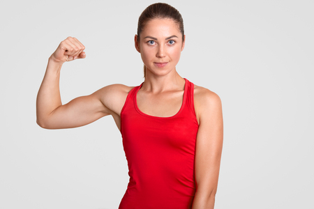 Horizontal shot of self determined young sportswoman in red t shirt, shows her muscles, has training in gym every day, stands against white background. People, wellbeing and lifestyle conceptの写真素材