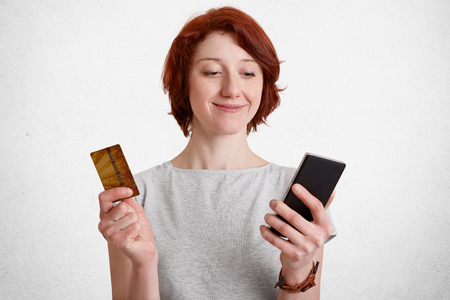 Happy delighted female with short hair, holds smart phone and plastic card, checks her bank account, being satisfied to recieve salary, uses online application, isolated over white concrete wallの写真素材