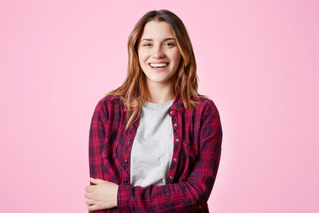 Positive female student rejoices to have holidays, dressed in checkered fashionable shirt, smiles happily isolated on pink backgroundの写真素材