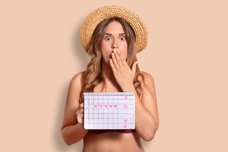 Shocked terrified female covers mouth with palm, shocked to have periods, holds period calendar,...