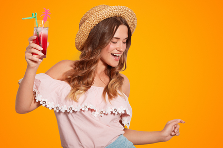 Joyful young European female in summer straw hat, wears fashionable blouse, holds cold fresh cocktail, being in good mood, isolated over orange background.の写真素材