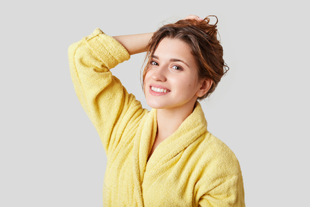 Pleased delightful young female model being in good mood after taking bath, dressed in casual yellow bathrobe, isolated over white background.の写真素材