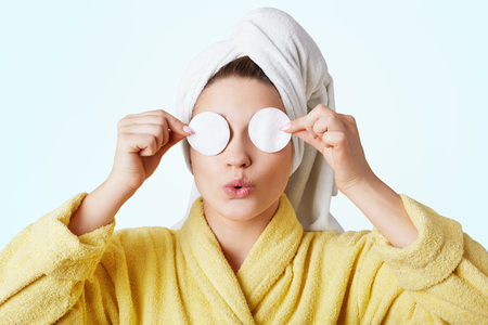 Funny woman foolishness after taking shower, wears towel and bathrobe, covers eyes with cotton wheels, going to apply cosmetics and do make up, isolated on white background.の写真素材