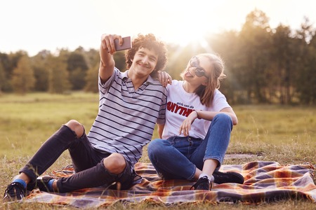 Cheerful girl and boy make selfie portrait of themselves, hold modern smart phone, sit on plaid on green field, dressed in summer clothing. Female and male youngsters use modern technology outsideの写真素材