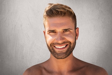 Positive young macho with stylish hairdo, hazel eyes, bristle and pleasant smile, demonstrating his white perfect teeth standing topless against grey concrete wall. Man with attractive appearanceの写真素材