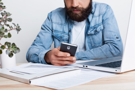 A film effect. Portrait of bearded man in jean shirt hoding smartphone in his hand while working with documents in office with cosy interior. Cropped portrait of succesful businessman using mobile.の写真素材
