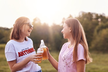 Best female friends toast with bottles of cold beer, have fun together, spend free time outdoor, spend summer holidays in countryside, laugh joyfully, enjoy relaxation. People and friendship conceptの写真素材