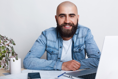 A happy young bald businessman having thick dark beard dressed in jean shirt siiting at the table in his office with laptop and smartphone being busy with documents. People, business and  technology.の写真素材