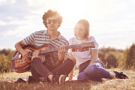Stylish male teenager with curly hair, wears sunglasses, holds music instrument, plays acoustic guitar, spends free time with girlfriend who has happy expression, enjoy good weather outdoor.の写真素材