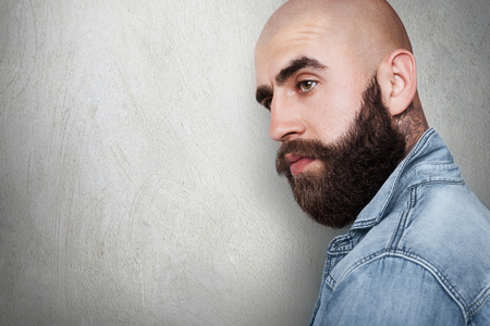 A sideways portraitwith copy space of fashionable bald hipster having tattoo on his neck, thick black eyebrows and beard, big dark eyes wearing jean shirt looking aside. People and lifestyle.の写真素材