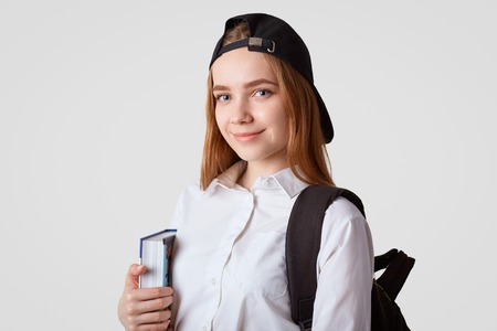 Blue eyed delighted schoolgirl holds book, carries backpack, wears black cap, going to library, reads and cramms material for exam, poses against white background. Studying and school conceptの写真素材