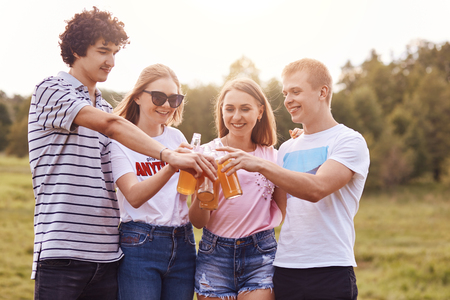 Photo of happy teenagers celebrate something or just have rest outdoor, clink bottles of beer, have delighted expressions, wear summer clothes, have fun together. People, picnic, lifestyle conceptの写真素材