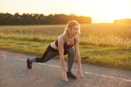 Attractive female wearing sport shoes and clothes, doing sport exercises on road against sunset,...