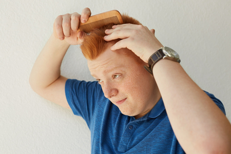 A redhead boy with freckles dressed in blue casual shirt brushing hair with comb on white background. Stylish handsome man combing his hair. Beauty and people concept.の写真素材