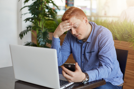 People, business, modern technology and communication concept.A young successful man with stylish red hair dressed in checked shirt sitting at cafe table, working on his laptop and messaging via phoneの写真素材