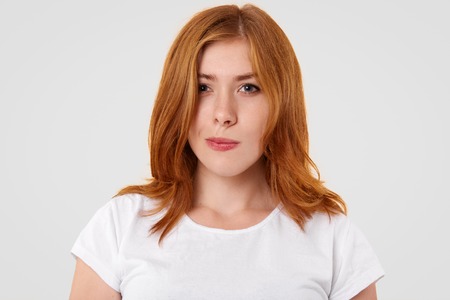 Doubtful pretty female purses lips and looks with hesitation, has reddish hair, dressed in casual t...