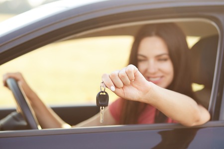 Happy young brunette female holds key while sits in luxury automobile, glad to recieve expensive...