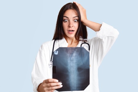 Terrific female doctor in white robe examines X ray picture of spine, holds hand on head, realizes that patient has scoliosis, isolated over light blue background. Woman practitioner indoor.の写真素材