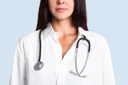 Cropped image of brunette young female therapist in white gown with phonendoscope, stands indoor, going to diagnose patients who came on dosctor`s appointement. People, medicine, health care conceptの写真素材