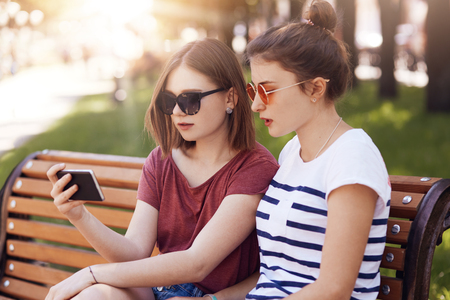Horizontal shot of pretty two female students in casual summer clothes, look attentivwly into mobile phone, watch intersting serial, recreat on bench while wait for someone. People, park, restの写真素材