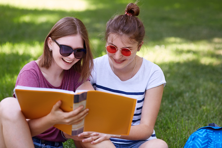 Shot of two cheerful friends have joyful looks into book, read something funny, wear sunglasses, pose against green grass background, spend time after classes in park. Youth and studying conceptの写真素材