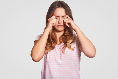 Photo of displeased young girl rubs eyes, being in low spirit, wears casual domestic clothes, poses against white background. Woman had bad sleep at night. Bedding concept. People and tirednessの写真素材