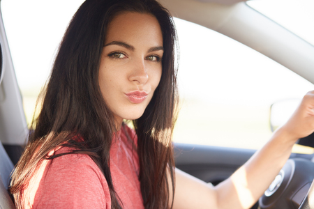 Close up shot of beautiful brunette girl pouts lips, has makeup, looks directly at camera, poses in automobile, keeps hands on wheel, makes grimace, flirts with boyfriend. Photo of female driverの写真素材