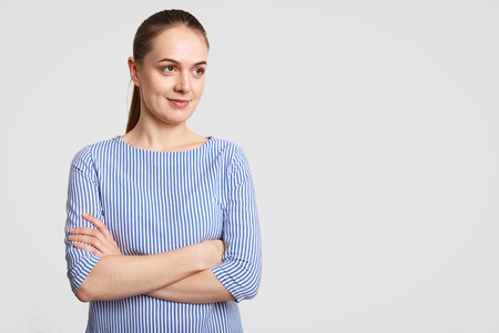 Horizontal shot of thoughtful European female with pony tail, keeps hands crossed, dressed in striped blouse, poses against white background with copy space aside for your promotion or advertisementの写真素材