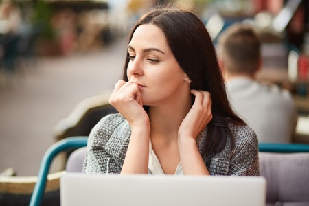 Thoughtful beautiful young lady looks aside with pensive expression, keeps hand on mouth, wrapped in warm coverlet, spends free time in outdoor cafe, works on course paper, uses laptop computerの写真素材