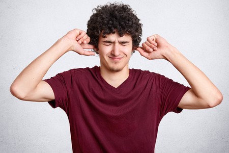 Horizontal shot of displeased man has curly hair, plugs ears, keeps eyes closed, doesnt want to hear loud noise, ignores somebody, wears loose t shirt, isolated over white background. Dont bother meの写真素材