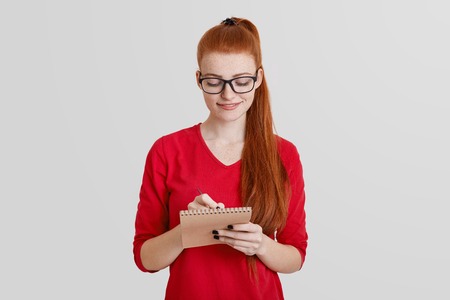 Pretty young woman with long ginger ponytail, writes notes in notebook, creates her reminder or working schedule, writes down information, isolated over white background. Female student studiesの写真素材