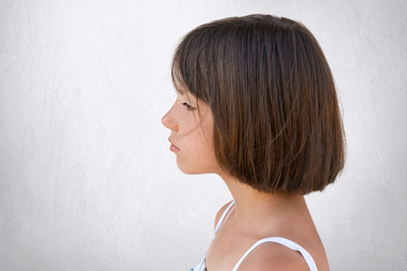 Sideways portrait of adorable freckled girl looking into distance while having dreamy expression isolated over white concrete wall. Little girl with short dark hair standing sideways with serious lookの写真素材