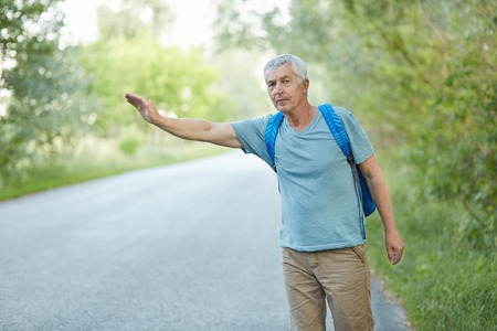 Outdoor shot of grey haired senior man in casual t shirt, has vacation trip, hitchhikes on road in countryside, has rucksack on back. Travelling, tourism and hitchhiking concept.の写真素材